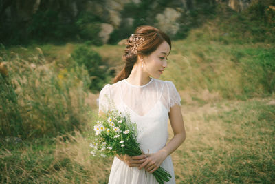 Bride holding flowers looking away while standing on grassy field