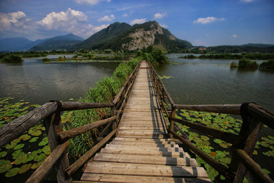 Wooden boardwalk amidst lake against mountains and sky on sunny day