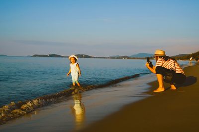 Mother photographing daughter on beach against sky