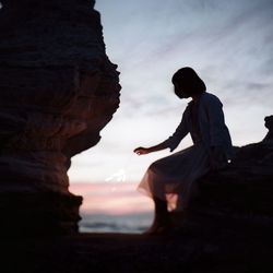 Full length of woman holding sparkle while sitting on rock against sky during sunset