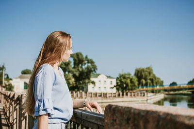 Lonely pensive young blonde woman in blue dress standing near the city river in summer sun 