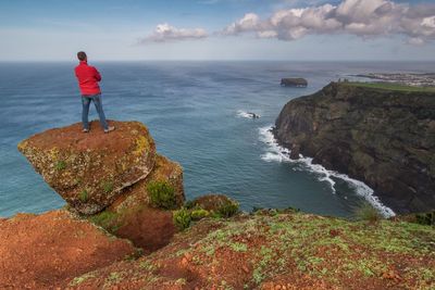 Rear view of man standing on cliff by sea against sky