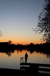 Rear view of silhouette man fishing while standing by lake during sunset