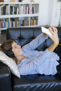 Smiling young woman using phone while lying on sofa at home