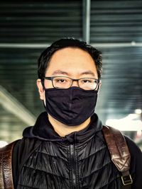 Portrait of young man wearing eyeglasses and black face mask against ceiling and lights.