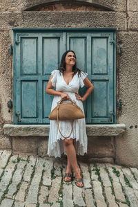 Full length portrait of beautiful young woman standing in front of blue window shutters in old town