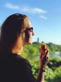 Side view of young woman eating ice cream cone while standing against blue sky