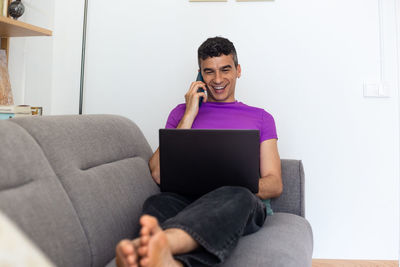 Full body of smiling transgender man using phone and laptop sitting on sofa at home