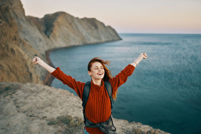 Smiling young woman with arms raised against sea against sky