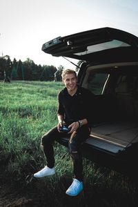 Young man smiling while sitting in car trunk on grassy field