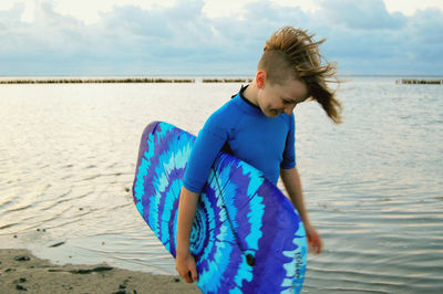 Boy with blue surfboard walking at beach against cloudy sky during sunset