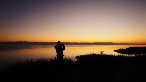 Silhouette woman photographing lake against sky during sunset