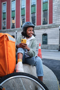 Positive african american female rider in helmet sitting near bicycle and thermo backpack while checking information on cellphone in city