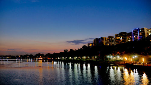 Illuminated buildings by river against sky at sunset