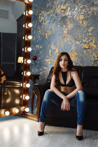 Portrait of young woman sitting on sofa by illuminated light bulbs