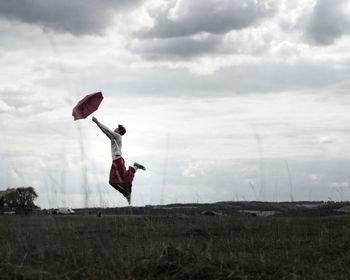 Side view of young man with umbrella jumping on grassy field against cloudy sky