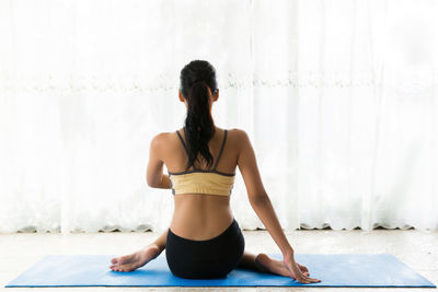 Rear view of young woman practicing yoga against curtain at home