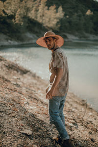 Portrait of young man wearing hat standing against lake in forest