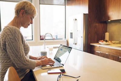 Side view of senior woman typing on laptop while sitting at kitchen table