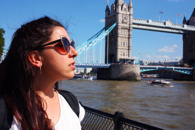 Beautiful young woman looking away while sitting on bridge against sky