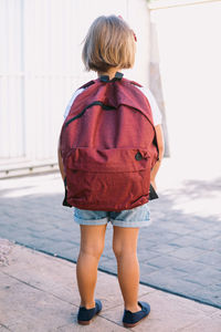 Back view of unrecognizable schoolchild with backpack standing on pavement in sunlight