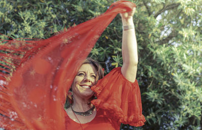 Smiling woman looking away while holding scarf against trees