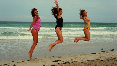 Happy friends jumping on sand against sea during sunset