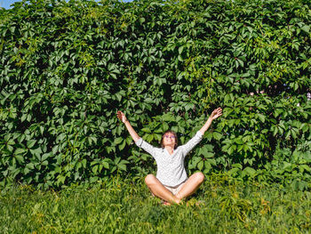 Wide smiling woman sits on grass lawn near wall all over covered with thick ivy. summer vibes.