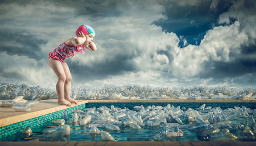 Side view of shocked girl looking at plastic bottles in swimming pool against cloudy sky