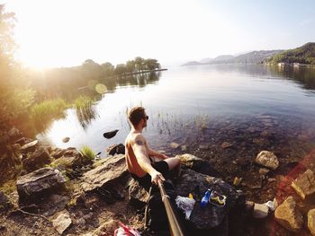 Shirtless man taking selfie using monopod while sitting on rock by lake