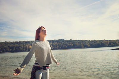 Woman with eyes closed standing by lake against cloudy sky