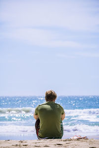 Rear view of man sitting on rock by sea against clear sky