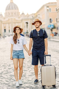 Happy young couple of travellers with hats and a suitcase hugging in the city and enjoying romance. 