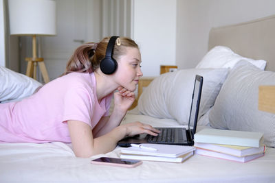 A young girl of 15-18 years old lies on a bed with a laptop and headphones and phone