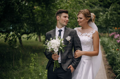 Newlywed couple looking at each other while standing against trees