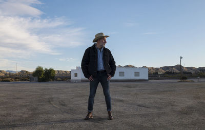 Adult man in cowboy hat standing in front of white building against sky