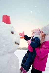 Teenage girl carrying sister by snowman against sky