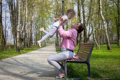 On a bench in a city park, a mother lifts her child high into the air.
