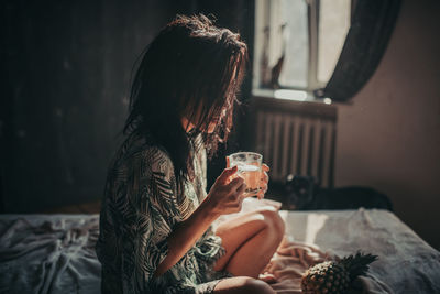 Midsection of woman drinking glass while sitting on bed at home