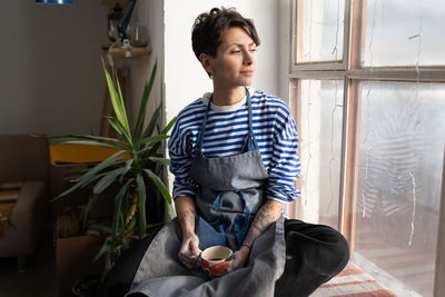 Optimistic italian woman artist or craftsman with tea cup sits on windowsill and looks out window