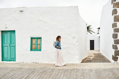 Side view of female with backpack walking on pavement against white houses and cloudy gray sky on town street in fuerteventura, spain