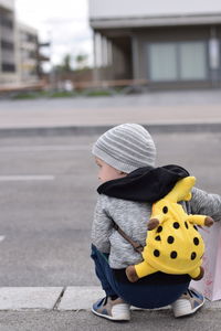 Rear view of boy with backpack crouching on street in city