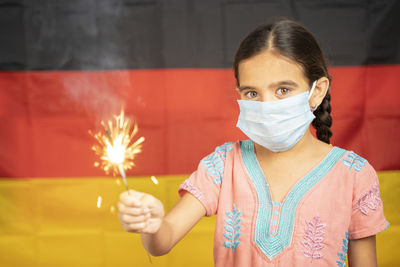 Portrait of girl wearing mask holding sparkler standing against german flag