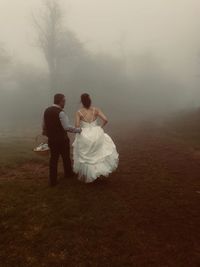 Rear view of bride and groom walking on field during foggy weather