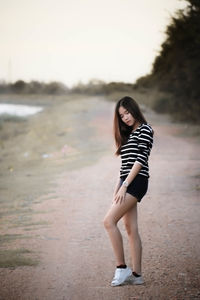 Side view of young woman standing on dirt road against clear sky