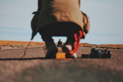 Man crouching on road in front of friend amidst field against sky