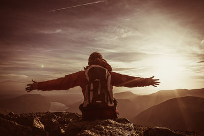 Rear view of hiker with backpack and arms outstretched leaning on rock against sky