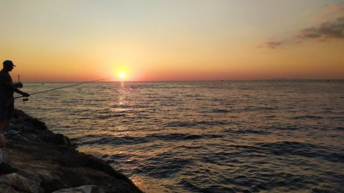 Silhouette man fishing on rocky shore against sky