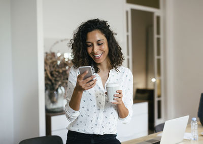Businesswoman in office using smart phone, holding cup of coffee