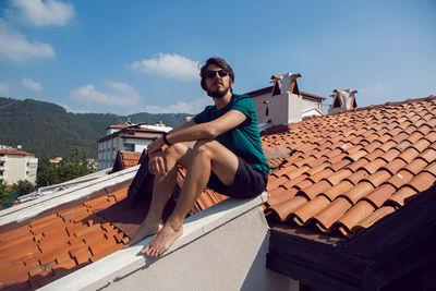 Man in the green t-shirt shorts and sunglasses sitting on the roof of a clay orange tile